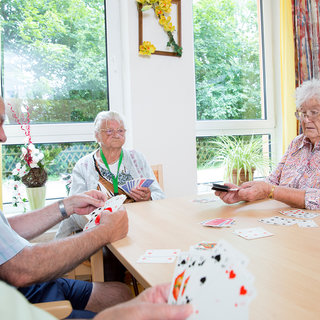 Vier Senioren sitzen an einem Tisch und spielen Karten. Zwei Frauen und zwei Männer sind konzentriert mit ihren Karten beschäftigt, während Spielkarten auf dem Tisch verteilt sind. Im Hintergrund sind Fenster mit viel Tageslicht und einige Pflanzen zu sehen.