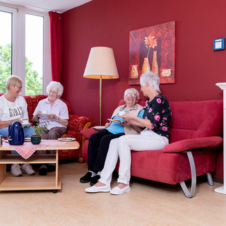 Vier ältere Frauen sitzen in einem gemütlichen Wohnzimmer. Zwei Frauen sind auf einer roten Couch, während die anderen beiden am Tisch sitzen. Sie wirken entspannt und unterhalten sich, umgeben von Pflanzen und einem warmen Raumlicht.