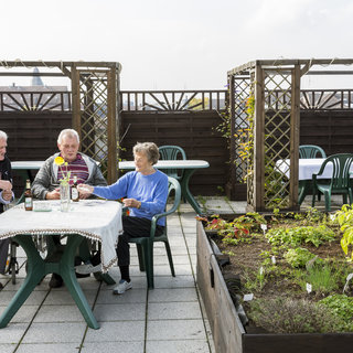 Drei ältere Menschen sitzen an einem Tisch auf einer Dachterrasse. Einer sitzt im Rollstuhl, während die anderen an einem Tisch stehen. Um sie herum gibt es Tische mit Stühlen und ein kleines Beet mit Pflanzen. Der Himmel ist teilweise bewölkt.