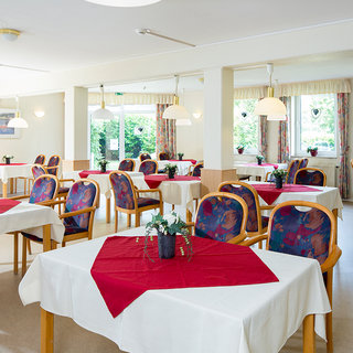 Ein helles Restaurant mit mehreren Tischen, die mit weißen Tischdecken und roten Tischläufern dekoriert sind. Die Stühle sind mit bunten Stoffen bezogen. Fenster lassen Tageslicht hinein und der Raum wirkt freundlich und einladend. Kleine Blumenarrangements stehen auf den Tischen.
