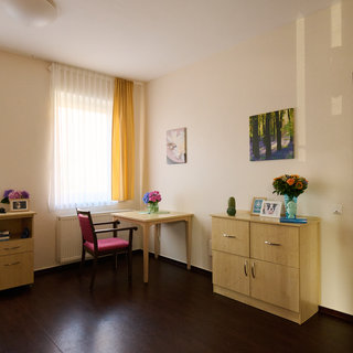 Das Bild zeigt ein helles Zimmer mit großen Fenstern, einem Tisch und einem Stuhl. An einer Wand stehen ein Bücherregal und ein Schrank. Auf dem Tisch befinden sich Blumen und Bilderrahmen. Die Wände sind in neutralen Farben gehalten, und der Boden ist dunkelbraun.