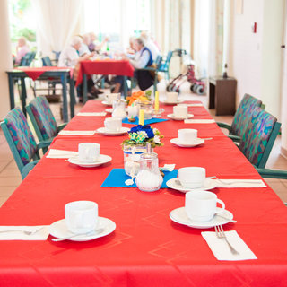 Ein gedeckter Tisch mit roten Tischdecken, dekoriert mit Blumen und Tellern. Auf dem Tisch stehen Kaffeetassen und Besteck. Im Hintergrund sind Menschen zu sehen, die am Tisch sitzen und sich unterhalten. Die Atmosphäre wirkt einladend und gesellig.