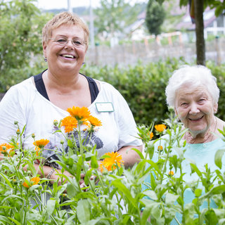 Zwei Frauen stehen lächelnd in einem Garten, umgeben von blühenden, orangefarbenen Blumen. Eine Frau, jünger mit kurzen, blonden Haaren, trägt ein helles T-Shirt, während die ältere Frau, mit weißem Haar, freundlich in die Kamera schaut. Beide strahlen Freude aus.