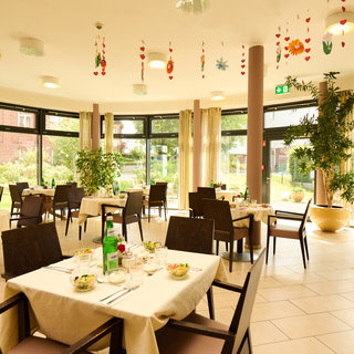 Ein helles Restaurant mit großen Fenstern, das eine grüne Gartenansicht bietet. Tische sind mit Tischdecken und Geschirr eingedeckt, Stühle umgeben die Tische. Pflanzen sind im Raum verteilt, und bunte Dekoration hängt von der Decke.