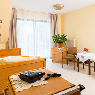 Ein helles Zimmer mit einem Krankenhausbett und einer Nachttischlampe. Neben dem Bett steht ein Telefon. Auf einem Tisch sind eine Blume und eine Pflanze. Außerdem gibt es einen Stuhl und eine Kommode. Große Fenster sorgen für viel Tageslicht.