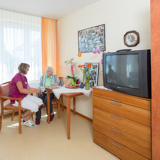 Zwei Frauen sitzen in einem hellen Raum. Eine ältere Dame mit grauen Haaren und einer grünen Bluse hört aufmerksam zu, während eine Betreuungskraft in lila Kleidungsstücken mit ihr spricht. Auf dem Tisch stehen Blumen und kleinere Snacks. Ein Fernseher und eine Kommode sind im Hintergrund sichtbar.