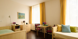 Das Bild zeigt ein helles Zimmer mit einem Bett, einem Tisch und zwei Stühlen. An der Wand hängt ein Gemälde, und auf dem Tisch steht eine Vase mit Blumen. Große Fenster sind mit gelben Vorhängen dekoriert, die Licht in den Raum lassen.