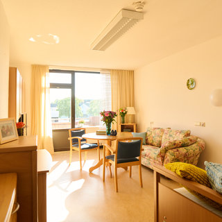 Ein helles Zimmer mit einem Fenster, einer gemütlichen Sitzecke mit einem Blumenmuster-Sofa, einem Esstisch und Stühlen. Auf dem Tisch stehen Blumenvasen. Es gibt einen Schrank und Fotos an der Wand, was eine einladende Atmosphäre schafft.