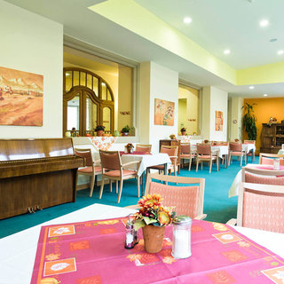 Ein Speisesaal mit hellen Wänden und freundlicher Atmosphäre. Tische mit rosa Tischdecken sind ordentlich gedeckt. Im Hintergrund steht ein Klavier, und an den Wänden hängen Bilder. Die Einrichtung ist modern und einladend, ideal für gesellige Mahlzeiten.