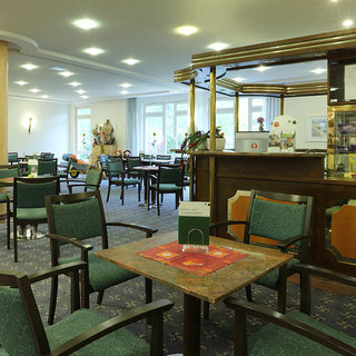 Ein einladender Raum mit mehreren Tischen und Stühlen ist zu sehen. Die Möbel sind in neutralen Farben gehalten, und der Raum ist hell erleuchtet. Ein Bereich mit einer Theke und dekorativen Elementen ist im Hintergrund sichtbar, was eine freundliche Atmosphäre schafft.