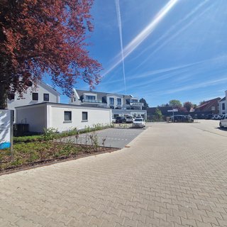 Das Bild zeigt einen sonnigen Außenbereich einer Einrichtung namens CURA. Es ist ein gepflasterter Parkplatz mit mehreren Fahrzeugen. Im Vordergrund befindet sich ein Schild, das Dienstleistungen wie Pflege und Wohnen anbietet. Im Hintergrund sind moderne Gebäude und ein strahlend blauer Himmel zu sehen.