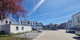 Das Bild zeigt einen sonnigen Außenbereich einer Einrichtung namens CURA. Es ist ein gepflasterter Parkplatz mit mehreren Fahrzeugen. Im Vordergrund befindet sich ein Schild, das Dienstleistungen wie Pflege und Wohnen anbietet. Im Hintergrund sind moderne Gebäude und ein strahlend blauer Himmel zu sehen.