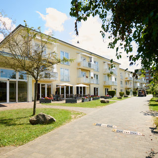 Ein modernes Wohngebäude mit mehreren Stockwerken, umgeben von einem gepflegten Garten. Der Eingang ist einladend mit Sitzgelegenheiten im Freien. Ein gepflasterter Weg führt entlang der Fassade, umgeben von Bäumen und Pflanzen. Die Sonne scheint, und der Himmel ist blauen.