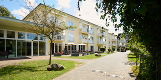 Ein modernes Wohngebäude mit mehreren Stockwerken, umgeben von einem gepflegten Garten. Der Eingang ist einladend mit Sitzgelegenheiten im Freien. Ein gepflasterter Weg führt entlang der Fassade, umgeben von Bäumen und Pflanzen. Die Sonne scheint, und der Himmel ist blauen.