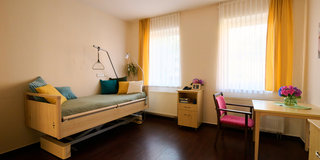 Ein helles Zimmer mit einem Krankenhausbett, das mit bunten Kissen dekoriert ist. Neben dem Bett steht eine Stehlampe und eine Pflanze. An einem Fenster hängen transparente Vorhänge. Am Tisch mit einer pinken Stuhl sind Blumen in einer Vase und ein Telefon.