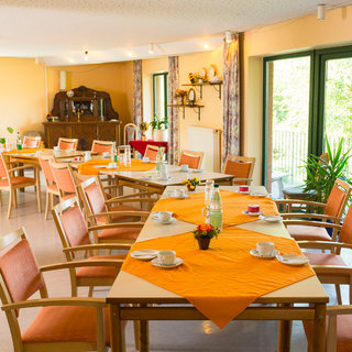 Ein hell erleuchteter Speisesaal mit mehreren Tischen, die mit orangenen Tischdecken gedeckt sind. Jede Tafel hat Tassen und Gläser, und Pflanzen sind im Raum verteilt. Der Raum bietet einen Blick auf die Natur durch große Fenster.