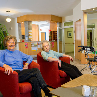 Zwei ältere Frauen sitzen in roten Sesseln in einem hellen Raum. Eine Frau lächelt, während die andere nachdenklich aussieht. Im Hintergrund sind Pflanzen und ein Tisch mit Geschirr sichtbar, was auf eine gesellige Umgebung hinweist.