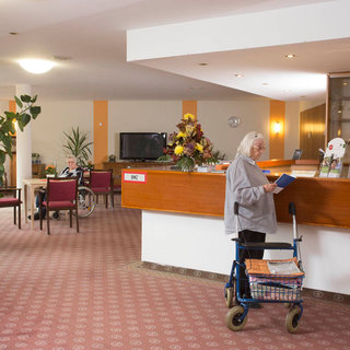 Eine ältere Frau steht an der Rezeption eines Eingangsbereichs. Sie hält ein Buch in der Hand und hat einen kleinen Einkaufstrolley bei sich. Im Hintergrund sind Sitzgelegenheiten und Pflanzen sichtbar, die Atmosphäre ist freundlich und einladend.