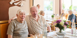 Zwei ältere Menschen sitzen zusammen an einem Tisch in einem hellen Raum. Sie lächeln und wirken freundlich. Im Hintergrund sind weitere ältere Personen und eine Blumenvase auf dem Tisch sichtbar, die eine angenehme Atmosphäre schaffen.