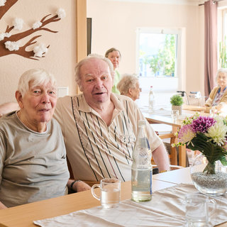 Zwei ältere Menschen sitzen zusammen an einem Tisch in einem hellen Raum. Sie lächeln und wirken freundlich. Im Hintergrund sind weitere ältere Personen und eine Blumenvase auf dem Tisch sichtbar, die eine angenehme Atmosphäre schaffen.