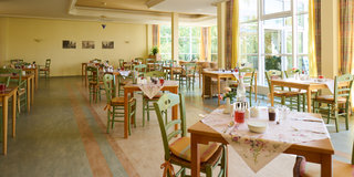 Ein hell erleuchteter Speisesaal mit mehreren Tischen und Stühlen. Die Tische sind mit Tischdecken, Tellern und Getränken gedeckt. Große Fenster lassen viel Tageslicht herein und bieten einen Blick auf den Außenbereich. Die Atmosphäre wirkt freundlich und einladend.