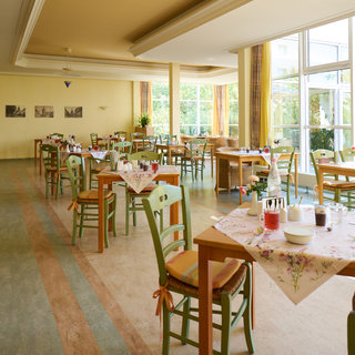 Ein hell erleuchteter Speisesaal mit mehreren Tischen und Stühlen. Die Tische sind mit Tischdecken, Tellern und Getränken gedeckt. Große Fenster lassen viel Tageslicht herein und bieten einen Blick auf den Außenbereich. Die Atmosphäre wirkt freundlich und einladend.