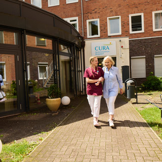 Zwei Frauen gehen auf dem Gehweg eines Pflegeheims. Eine der Frauen trägt eine Pflegeuniform und hält die andere, eine ältere Dame, am Arm. Im Hintergrund sind einige Fenster und eine grüne Rasenfläche zu sehen.