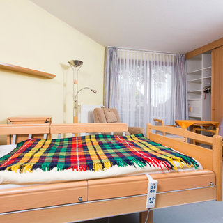 Ein gemütliches Zimmer mit einem elektrischen Bett, bedeckt mit einer karierten Decke. Im Hintergrund sieht man einen Sessel, einen Tisch und ein Fenster mit Vorhängen. Der Raum hat eine helle, freundliche Atmosphäre.