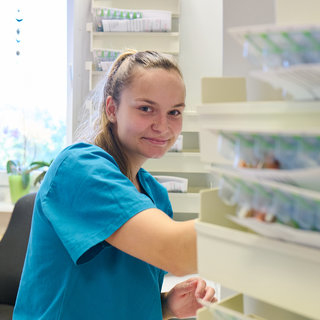 Eine junge Frau in einem blauen Arbeitshemd lächelt, während sie an einem Regal mit Medikamenten arbeitet. Im Hintergrund sind Pflanzen und ein Bürostuhl zu sehen, was auf eine freundliche Arbeitsumgebung hinweist.