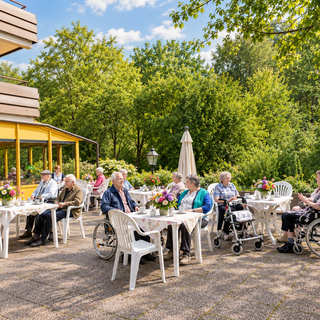 Ältere Menschen sitzen auf einer Terrasse vor einem Gebäude und genießen eine gemeinsame Zeit im Freien. Tische sind mit Blumen dekoriert, und die Umgebung ist grün mit Bäumen. Einige Personen sitzen in Rollstühlen, während andere auf Stühlen plaudern.