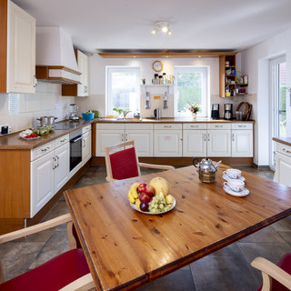 Eine helle, moderne Küche mit Holzoberflächen. Ein rechteckiger Tisch in der Mitte trägt eine Schale mit verschiedenen Früchten, darunter Äpfel, Trauben und eine Melone. Im Hintergrund sind Küchenschränke, ein Herd und Fenster, die Tageslicht hereinlassen.
