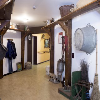 Der Flur ist von rustikalen Holzbalken gesäumt und zeigt verschiedene landwirtschaftliche Gegenstände sowie Kleidung. An den Wänden hängen Werkzeuge und Dekorationen, während der Boden aus hellem Material besteht. Der Raum vermittelt eine ländliche, einladende Atmosphäre.
